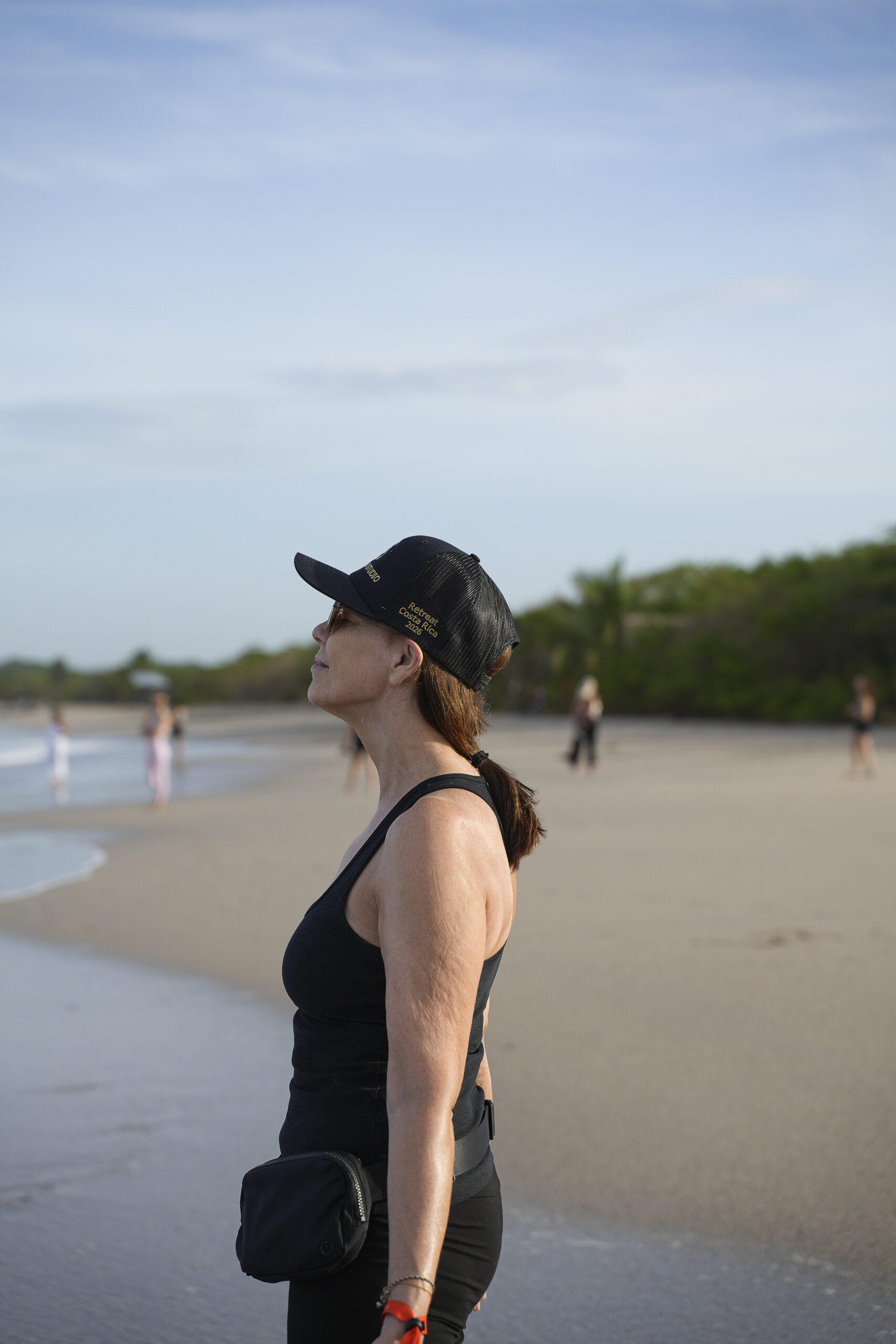 Side profile of a woman in a black cap and tank top standing on a beach, with a small black waist bag at her hip, looking toward the water.